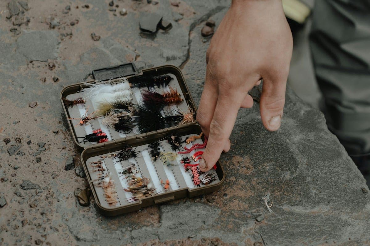 A hand pointing at an open fly box on a rock, its two halves lined with rows of flies in many sizes and colors