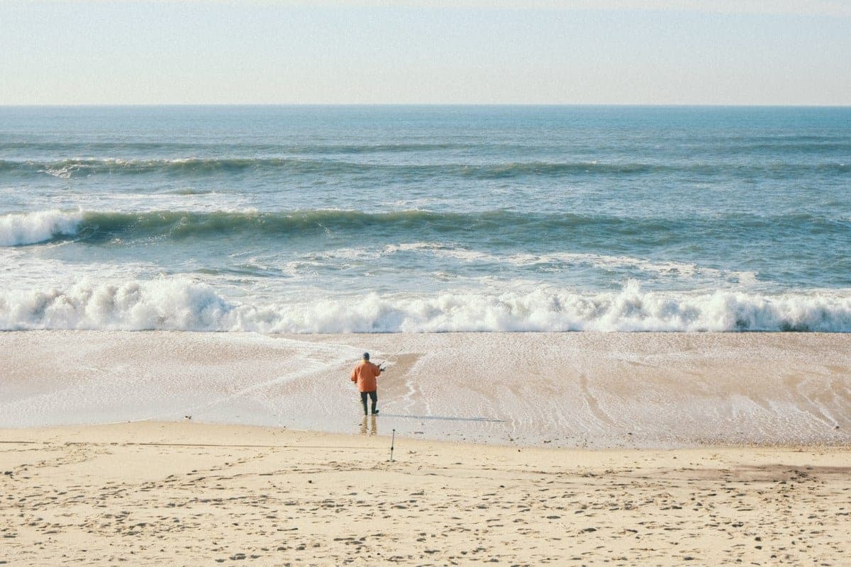 An angler fishing at the edge of the surf, with an empty sand spike pushed into the beach behind him